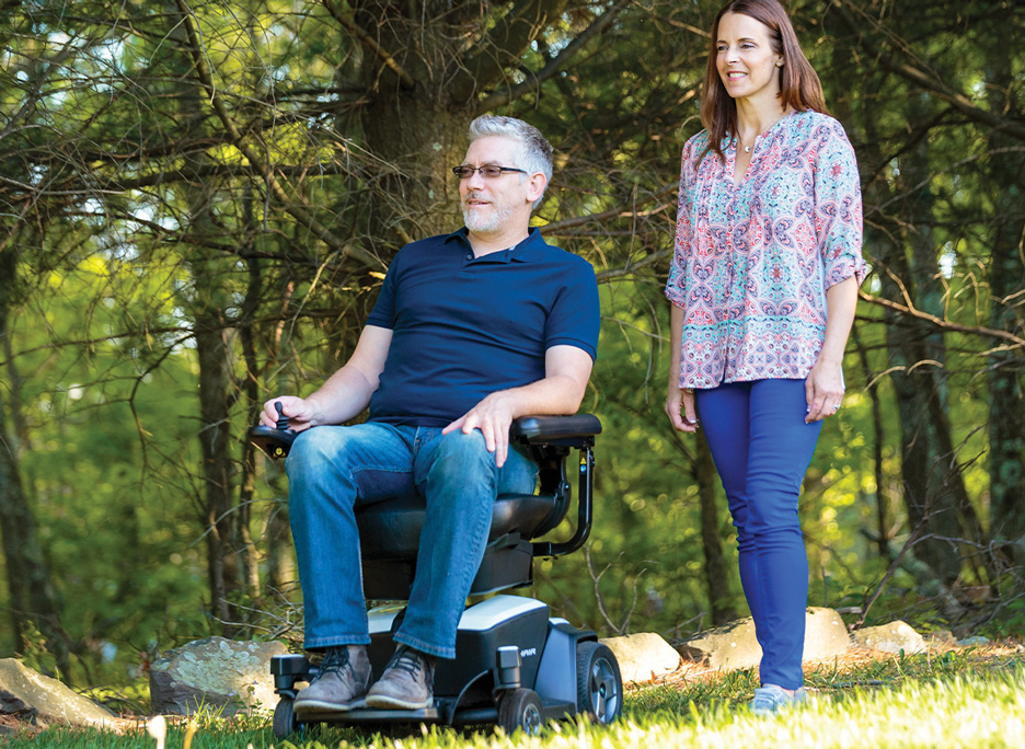 Person using power wheelchair on outdoor trail with person standing beside them